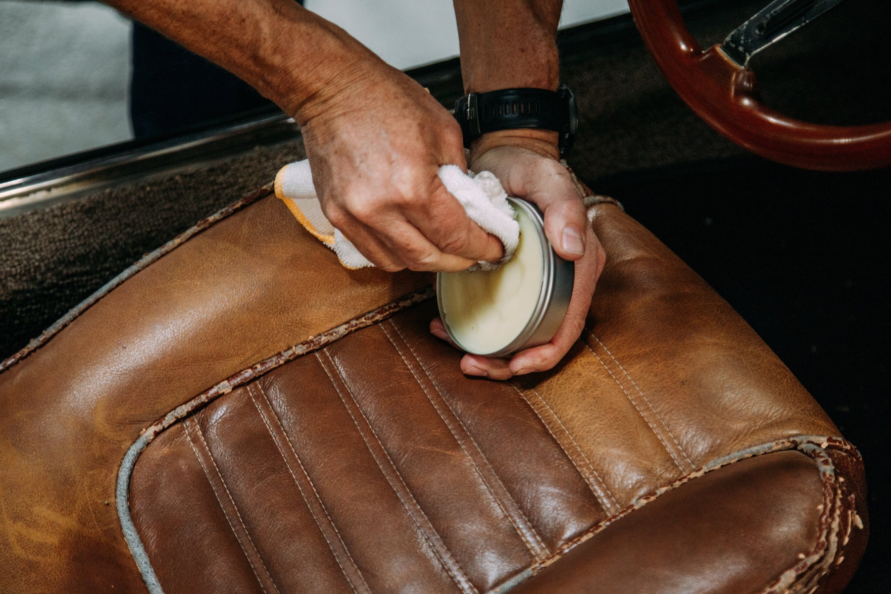 A man's hand holding a cloth, poised over an open can of 'Leather Love' leather conditioner, ready to rejuvenate faded leather seats